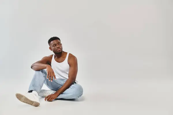 Junger Afroamerikaner sitzt mit gekreuzten Beinen im Studio vor grauem Hintergrund. — Stockfoto