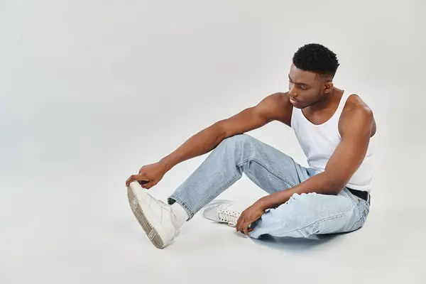 Um jovem e elegante afro-americano senta-se calmamente no chão em trajes casuais de jeans e um top de tanque. — Fotografia de Stock