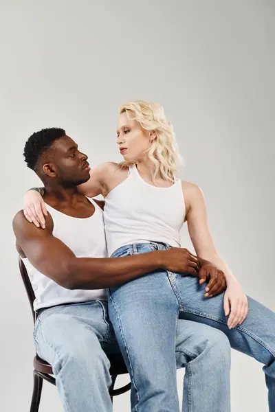 A young interracial couple leisurely seated on a chair in a studio against a grey background. — Stock Photo