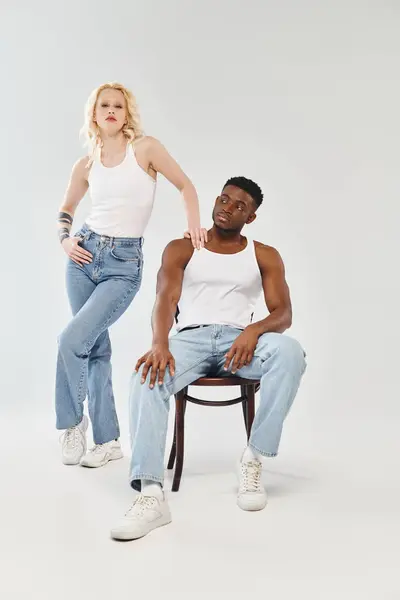 A young interracial couple sit on a chair in a studio against a grey background, exuding a sense of unity and togetherness. — Stock Photo
