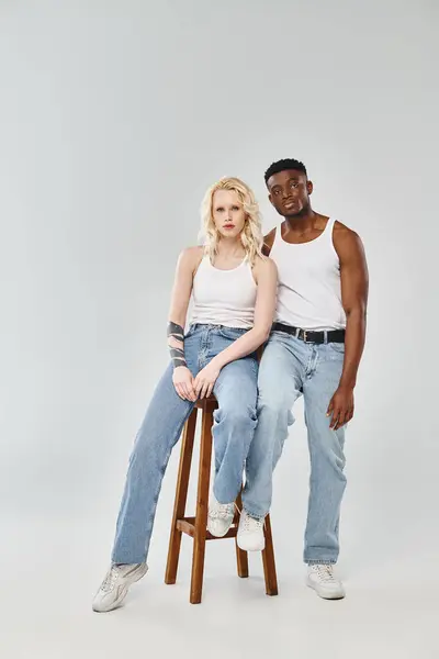 Um jovem casal interracial senta-se graciosamente junto em um banquinho em um estúdio contra um fundo cinza. — Fotografia de Stock