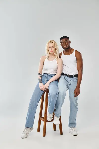 Un jeune couple interracial s'assoit gracieusement ensemble sur un tabouret dans un studio sur fond gris. — Photo de stock