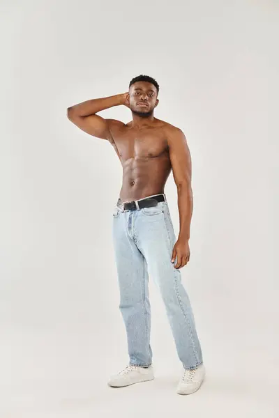 Un joven afroamericano sin camisa posa en un estudio sobre un fondo gris. - foto de stock