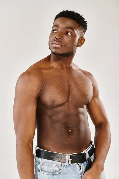 Un giovane uomo afroamericano senza maglietta posa con grazia in uno studio su uno sfondo grigio. — Foto stock