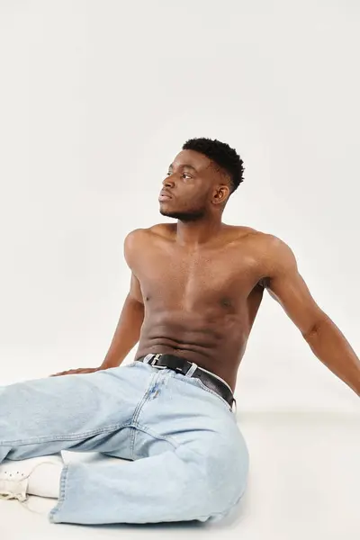 Без сорочки афроамериканський чоловік демонструє силу і впевненість, коли він сидить на землі в студії на сірому тлі. — стокове фото