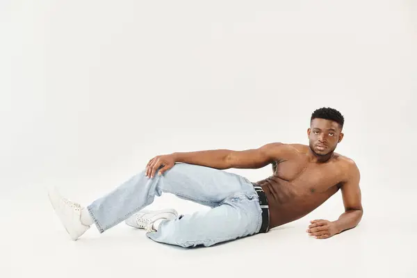 Um jovem afro-americano sem camisa faz uma pose cativante no chão em um estúdio contra um fundo cinza. — Fotografia de Stock