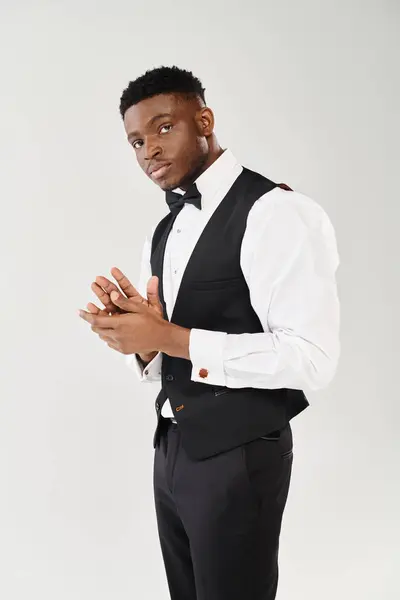 Um jovem afro-americano elegante fica confiante em um colete preto e camisa branca contra um fundo de estúdio cinza. — Fotografia de Stock
