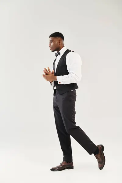 Un joven y guapo novio afroamericano camina elegantemente en un chaleco y corbata en un estudio sobre un fondo gris. — Stock Photo