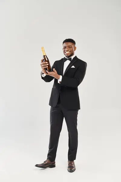 Le beau marié afro-américain en smoking tient une bouteille de champagne, prêt pour un toast de fête. — Photo de stock