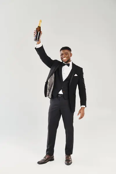 Guapo novio afroamericano en esmoquin sosteniendo una botella de champán, exudando elegancia y encanto en un ambiente de estudio. - foto de stock