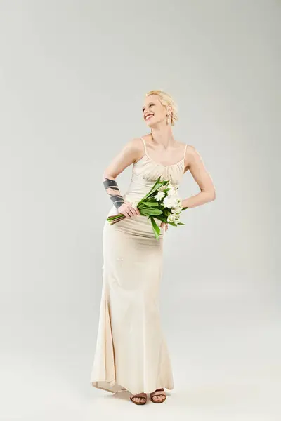 Una novia rubia impresionante en un vestido de novia con gracia la celebración de un vibrante ramo de flores sobre un fondo gris. - foto de stock