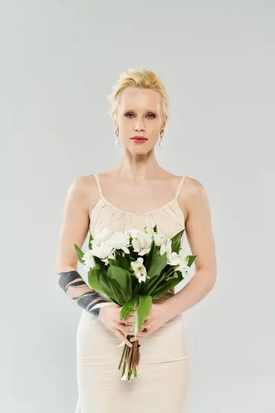 Ethereal blonde bride in white dress, gracefully holding vibrant bouquet of flowers against a grey backdrop. — Stock Photo