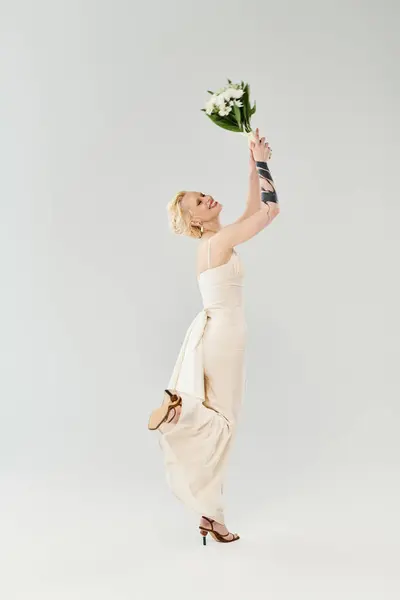 A beautiful blonde bride in a white dress delicately holds a bouquet of flowers in a studio against a grey background. — Stock Photo