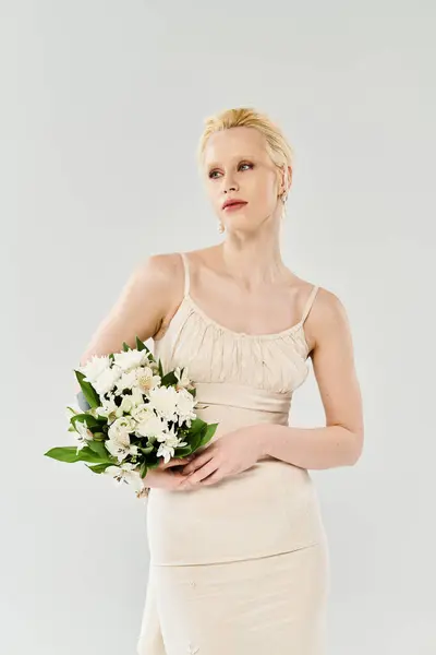 Une belle mariée blonde en robe tient un bouquet de fleurs dans un studio sur fond gris. — Photo de stock