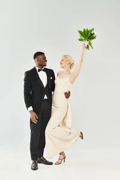 Une belle mariée blonde et marié afro-américain, habillé en tenue formelle, tenant un bouquet de fleurs sur un fond gris. — Photo de stock