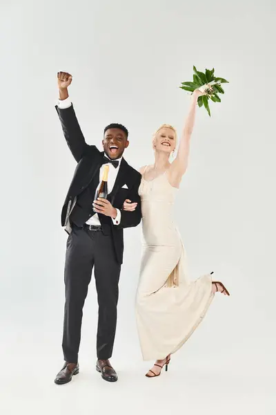 A beautiful blonde bride in a wedding dress and an African American groom holding flowers in a studio on a grey background. — Stock Photo