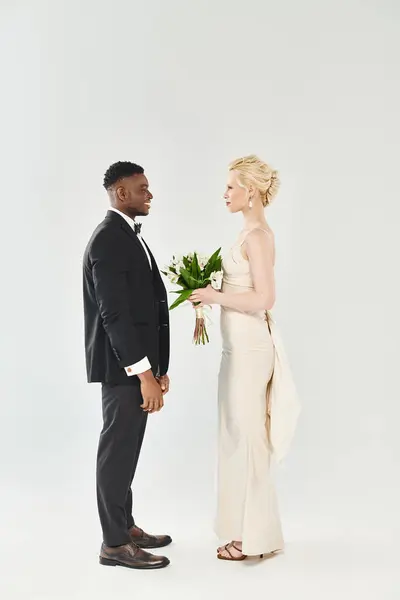 Una hermosa novia rubia en un vestido de novia está junto a un novio afroamericano en un estudio sobre un fondo gris. - foto de stock
