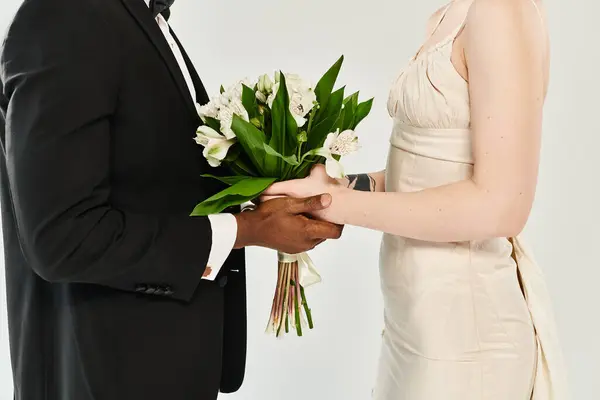 Uno sposo afroamericano in smoking tiene un mazzo di fiori accanto a una bella sposa bionda nel suo abito da sposa. — Foto stock
