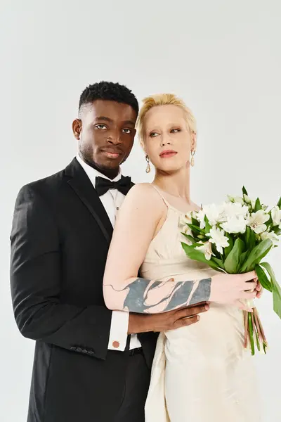 Une belle mariée blonde en robe de mariée et un marié afro-américain en smoking tenant un bouquet sur un fond gris. — Photo de stock