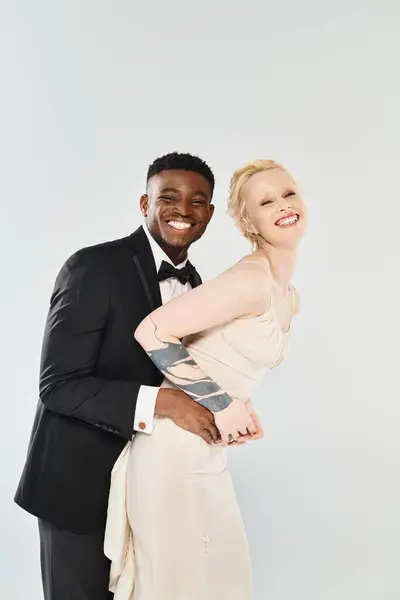 A beautiful blonde bride in a wedding dress and African American groom in a tuxedo pose together in a studio against a grey background. — Stock Photo