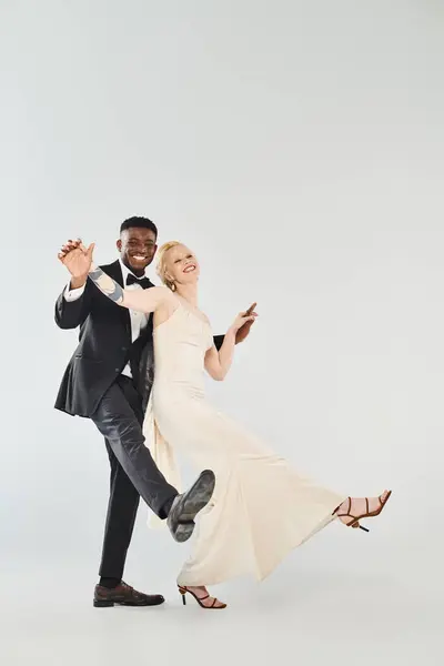 Una hermosa novia rubia en un vestido de novia y novio afroamericano bailando con gracia juntos en un estudio sobre un fondo gris. - foto de stock