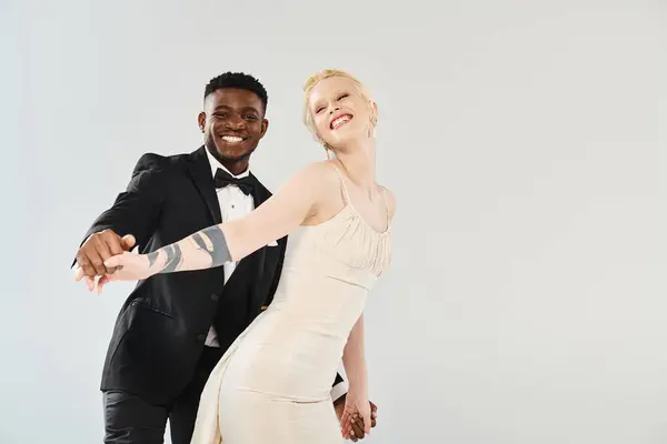 Eine schöne blonde Braut im Hochzeitskleid steht neben einem afroamerikanischen Bräutigam im scharfen Smoking vor grauem Hintergrund.. — Stockfoto