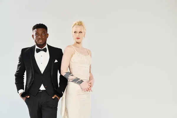 An African American man in a tuxedo stands next to a beautiful blonde woman in a white dress in a studio against a grey background. — Stock Photo