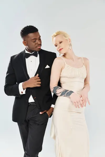 An African American groom in a tuxedo and a beautiful blonde bride in a wedding dress standing together in a studio on a grey background. — Stock Photo