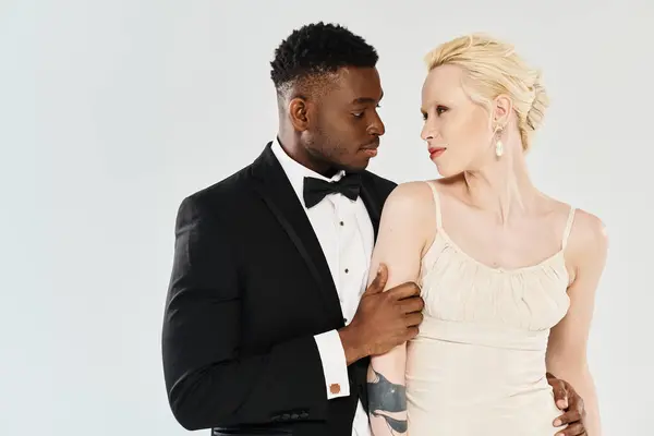 Красива блондинка в білій весільній сукні стоїть поруч з афроамериканським нареченим в класичному смокінгу, обидві в студії на сірому фоні. — стокове фото