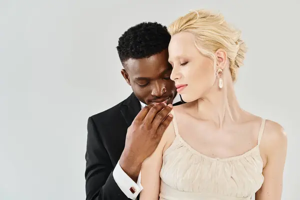 Una hermosa novia rubia en un vestido de novia está junto a su novio afroamericano en un estudio sobre un fondo gris. - foto de stock