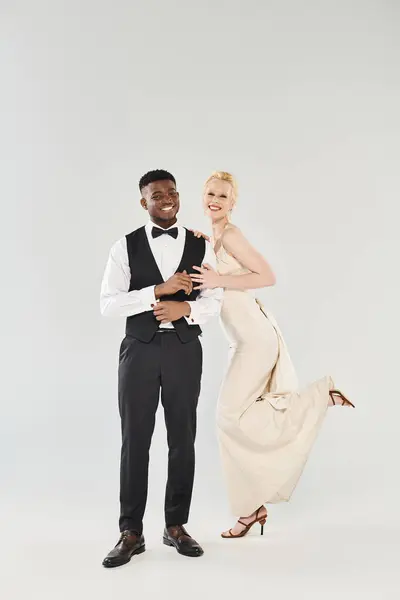 A beautiful blonde bride in a wedding dress and an African American groom in a tuxedo pose together in a studio against a grey background. — Stock Photo