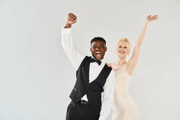 A beautiful blonde bride in a wedding dress and an African American groom pose elegantly in a studio against a grey background. — Stock Photo