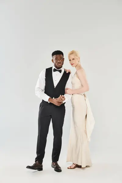 Uno sposo afroamericano in smoking e una bella sposa bionda in un abito da sposa fluente posano elegantemente in un ambiente da studio. — Foto stock