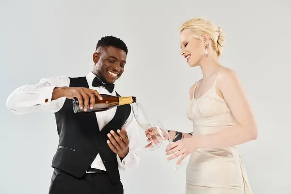 Un homme en smoking verse du champagne dans une main de femme, pendant qu'ils célèbrent dans un cadre de studio avec une belle mariée blonde dans une robe de mariée et un marié afro-américain sur un fond gris. — Photo de stock