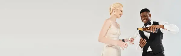 Une belle mariée blonde dans une robe de mariée se tient à côté d'un marié afro-américain dans un smoking sur fond de studio gris. — Photo de stock