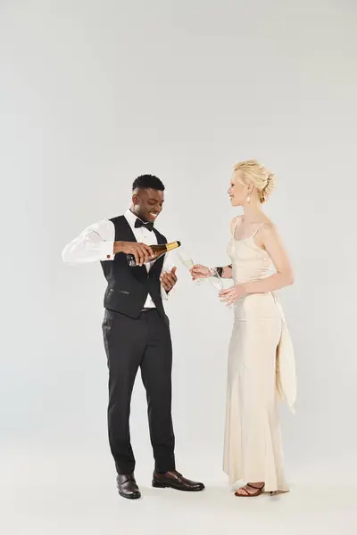 Une belle mariée blonde dans une robe de mariée et un marié afro-américain debout l'un à côté de l'autre dans un studio sur un fond gris. — Photo de stock