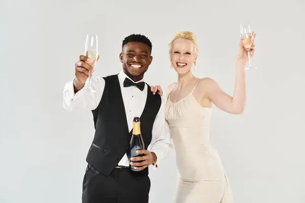 A beautiful blonde bride in a wedding dress and African American groom holding champagne glasses in a studio on a grey background. — Stock Photo