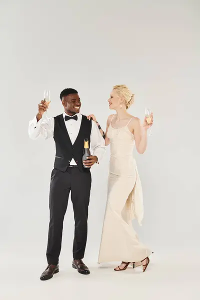 Une belle mariée blonde en robe de mariée et marié afro-américain tenant des flûtes à champagne dans un studio sur fond gris. — Photo de stock