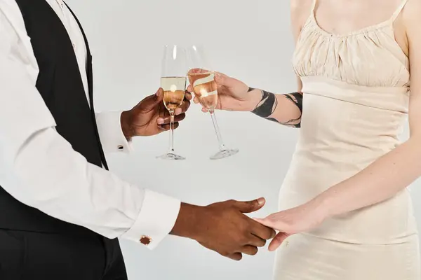 Una hermosa novia rubia con un vestido de novia y un novio afroamericano sosteniendo copas de champán en un estudio sobre un fondo gris. - foto de stock