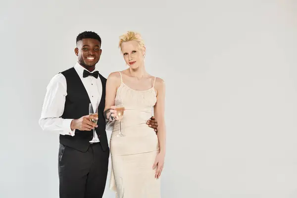 Una hermosa novia rubia en un vestido de novia y un novio afroamericano en esmoquin posan elegantemente en un estudio sobre un fondo gris. - foto de stock