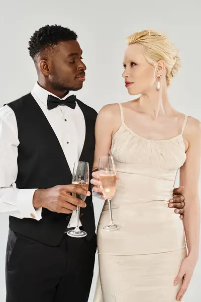 A beautiful blonde bride and African American groom standing together, holding wine glasses in a studio on a grey background. — Stock Photo