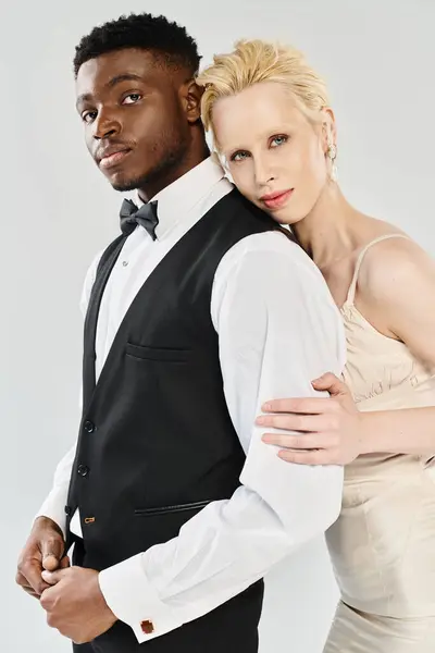 A beautiful blonde bride in a wedding dress and an African American groom in a tuxedo stand in a studio against a grey background. — Stock Photo