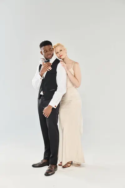 A beautiful blonde bride in a wedding dress and an African American groom stand next to each other in a studio on a grey background. — Stock Photo