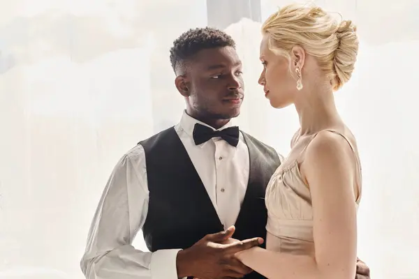 A beautiful blonde bride in a wedding dress and an African American groom standing together in a studio against a grey background. — Stock Photo