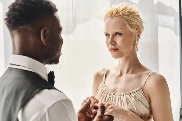 A beautiful blonde bride in a wedding dress and an African American groom stand side by side in a studio against a grey background. — Stock Photo