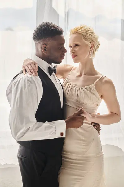 Eine schöne blonde Braut im Brautkleid und ein afroamerikanischer Bräutigam stehen nebeneinander in einem Studio auf grauem Hintergrund. — Stockfoto