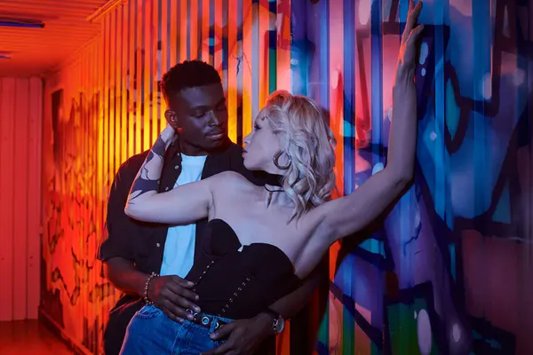 A blonde woman and African American man stand in front of a colorful wall covered in graffiti on an urban street. — Stock Photo