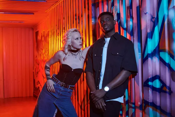 A blonde woman and an African American man stand side by side on an urban street adorned with colorful graffiti walls. — Stock Photo