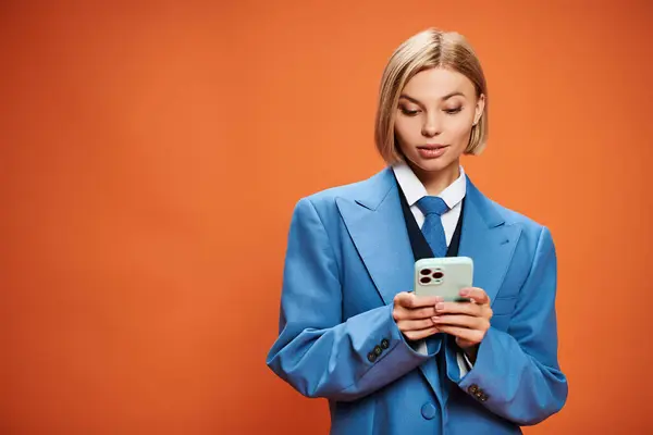 Mujer joven positiva con el pelo corto rubia posando con su teléfono inteligente sobre fondo naranja - foto de stock