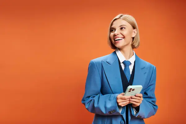 Alegre mujer bien vestida con pelo corto y rubio en traje elegante sosteniendo el teléfono sobre fondo naranja - foto de stock